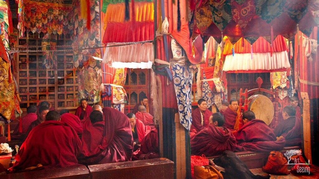 Monks in red robes gathered inside a Tibetan monastery hung with bright silk banners